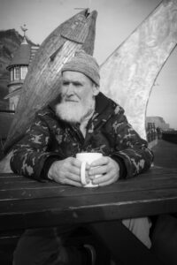 Man with bushy grey beard in Scarborough drinking tea in front of fish sculpture