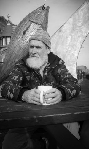Man with bushy grey beard in Scarborough drinking tea in front of fish sculpture